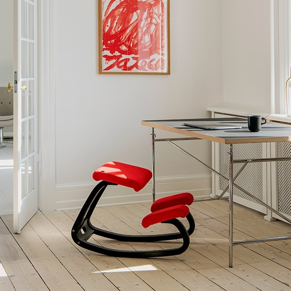 Rode ergonomische kniestoel Varier Variable Balans met zwart frame geplaatst bij een bureau in een lichte kamer