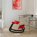 Rode ergonomische kniestoel Varier Variable Balans met zwart frame geplaatst bij een bureau in een lichte kamer